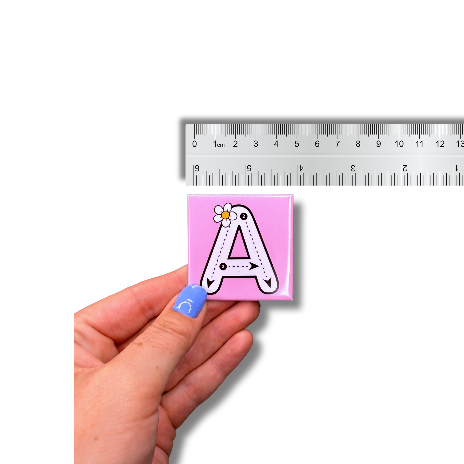 A hand holding a pink, 6 cm (approx 2.36 inch) square alphabet tracing magnet with the letter A, dashed tracing lines, and a small flower illustration for early learning practice.