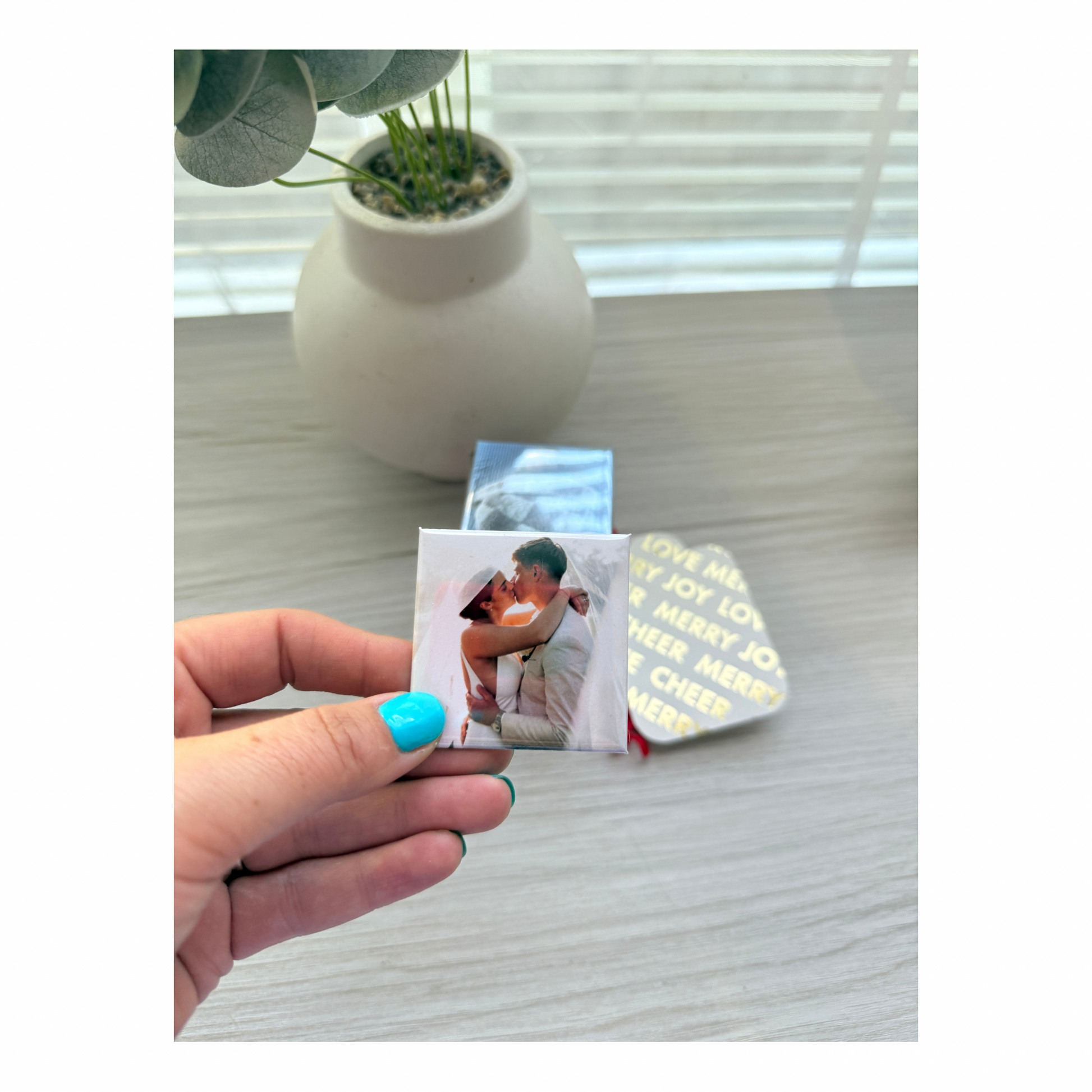 A hand holding a custom photo magnet next to a small plant and other magnets for lifestyle and size comparison.